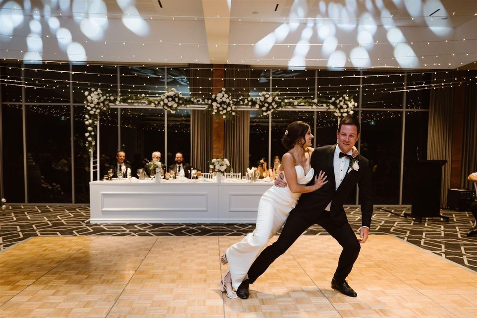 A bride and groom dance joyfully on a decorated ballroom floor, with guests seated at a long table under twinkling lights and floral arches.