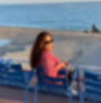 A relaxed traveler sitting comfortably on an iconic blue chair (La Chaise Bleue) along the Promenade des Anglais in Nice, France, enjoying a peaceful moment on the French Riviera with the Mediterranean Sea in the background.