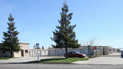 Single-story self-storage facility with gated vehicle entrance, metal unit doors, perimeter fencing, and a small office building fronting a suburban street.