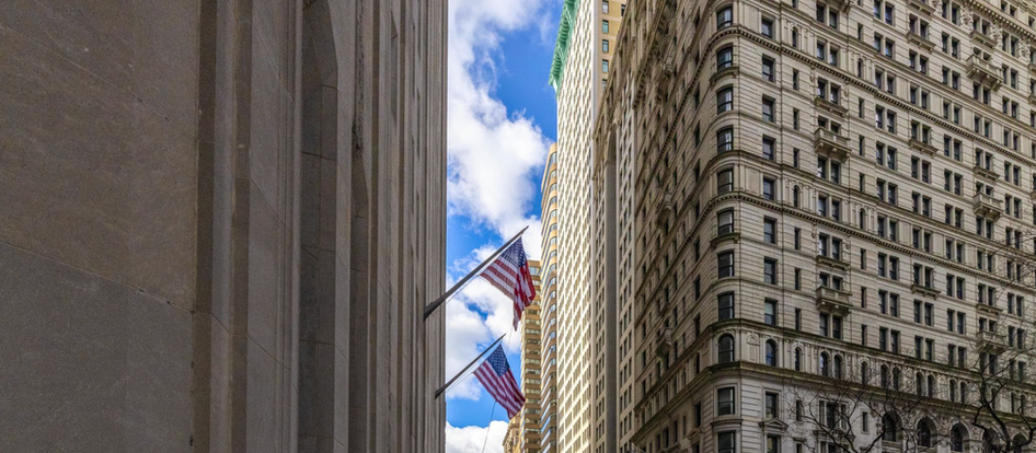 Dense urban commercial building facades along a Lower Manhattan street canyon, representing the concentration of commercial real estate assets in coastal markets facing escalating climate overlay risk designations.