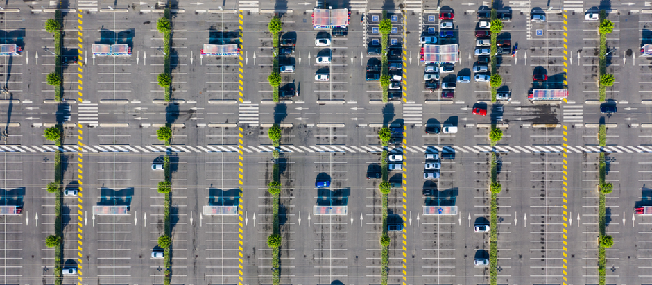 Aerial view of a large parking lot with scattered cars, tree-lined medians, covered cart stations, and marked crosswalks. Most cars are clustered in the upper right area.
