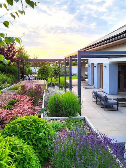 Widok na strefę relaksu, czyli nowoczesny, jasny taras wyłożony płytami betonowymi, na szare sofy tarasowe i metalową pergolę. Obok rozciąga się wielopoziomowy ogród z kwitnącymi bylinami i kolorowymi krzewami.