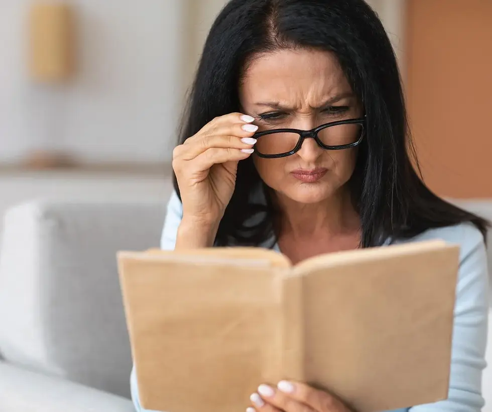 Woman with glasses reading a book, frowning in concentration. She's in a softly lit room with beige and white colors in the background.