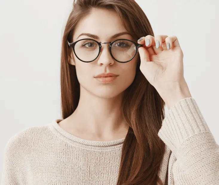Woman with long brown hair wearing glasses and a beige sweater adjusts her frames. Neutral expression, plain white background.