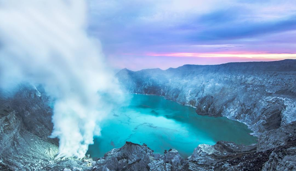 Steaming turquoise crater lake amid rocky gray landscape at dawn, with clouds of rising mist and a purple-pink sky in the background.