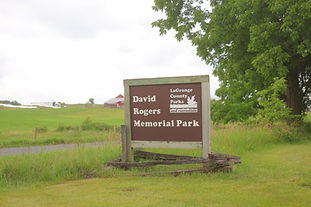Wide view of David Rogers Memorial Park with wood buildings and green trees.