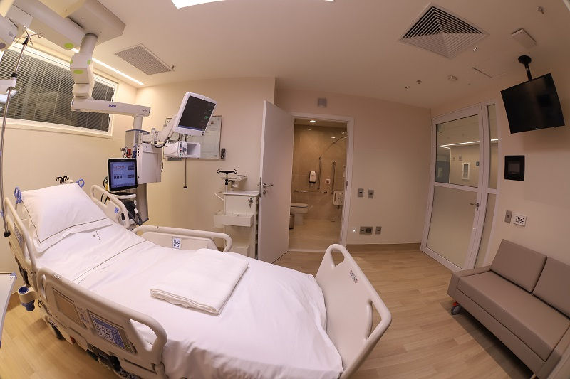 Suíte hospitalar com leito, televisão na parede, sofá à frente da cama e porta de vidro