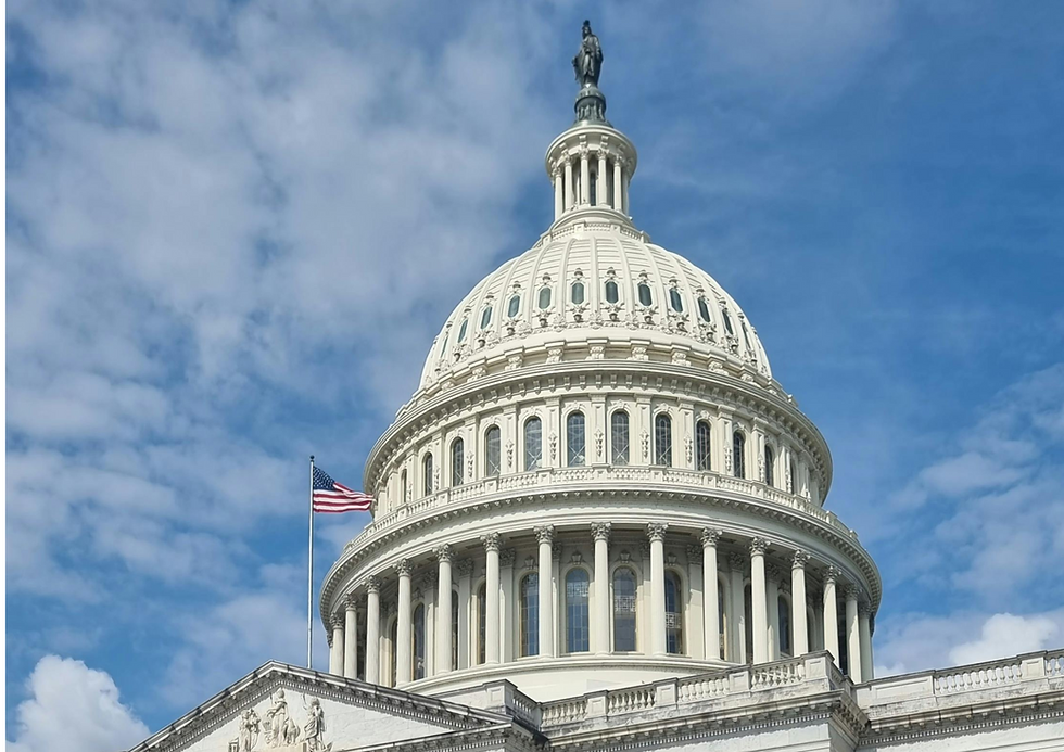 A detailed shot of the United States Capitol Building's dome, with the American flag visible on the left side of the frame and a blue sky with scattered clouds in the background.