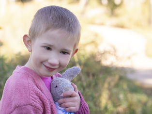 Smiling child in pink sweater holds a gray stuffed rabbit. Soft-focus background with greenery and sunlight. Warm, joyful mood.
