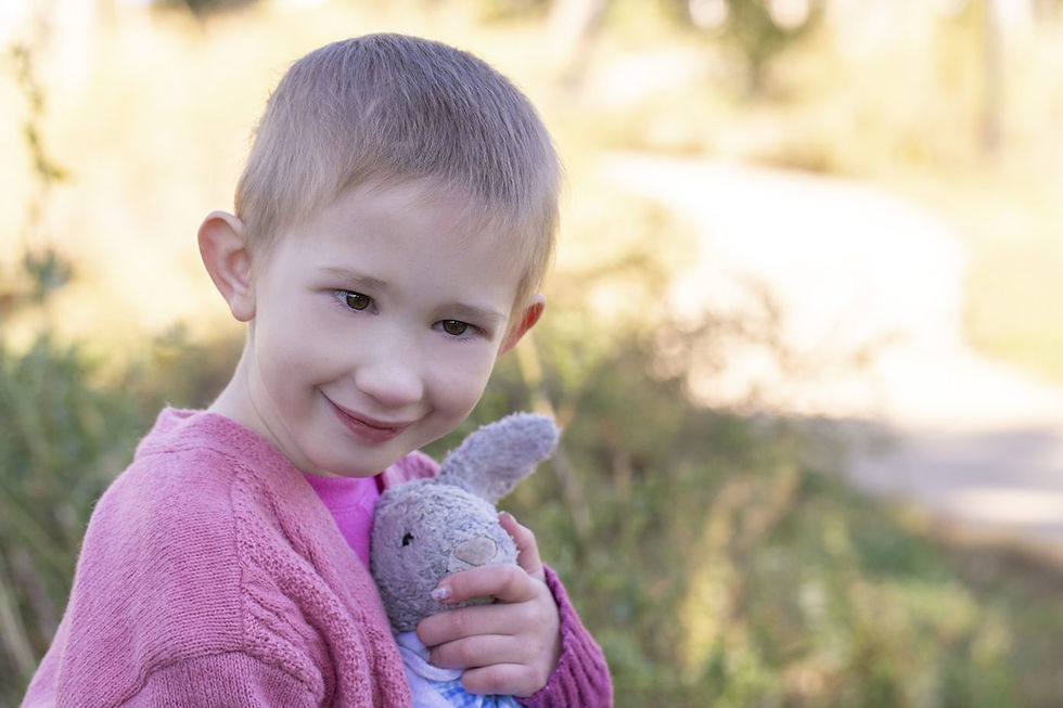Smiling child in pink sweater holds a gray stuffed rabbit. Soft-focus background with greenery and sunlight. Warm, joyful mood.
