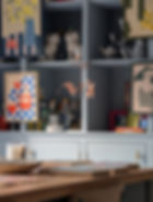 Art-filled blue cabinets at Emily Hadley gallery for Devon Open Studios with colourful paintings, ceramic dogs, bottles, and a wooden table holding frames and a candle. Warm, creative atmosphere.