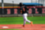 Normal Community's Max Heineman rounds the bases in a black and orange jersey atop a turf infield on a sunny day.