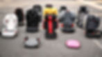 Eight colorful car seats and a pink bag are arranged on asphalt. Seats are in various styles and colors, with visible brand logos.