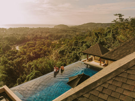 Two people relax in an infinity pool overlooking lush greenery and a coastal view at sunset. A villa with slate roofing is nearby.