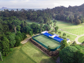 Aerial view of tennis and padel courts within a lush green park surrounded by dense trees. Bright blue and green colors dominate the peaceful setting.