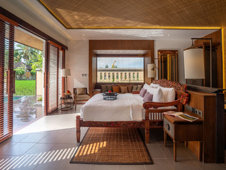 Luxurious bedroom with wooden decor, large bed, and cozy seating area. Sunlit view of a garden and pool through open blinds. Peaceful ambiance.