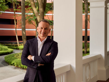 Man in a dark suit leaning against a white railing with arms crossed, smiling. Background shows trees and red building. Bright, outdoor setting.