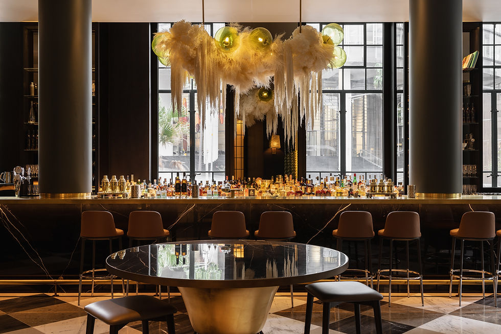 Chic bar with empty stools, ornate white chandelier, and bottles lined up. Marble floor, modern decor, large windows in the background.