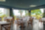 Elegant restaurant with wooden chairs and white tablecloths, set for dining. Open windows reveal lush greenery, creating a serene ambiance.