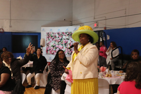 Woman in yellow dress speaking at 2024 Women's Tea event with microphone BGCCA.