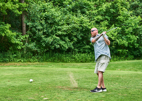 Man swinging golf club on green, ball in motion with a background of trees.