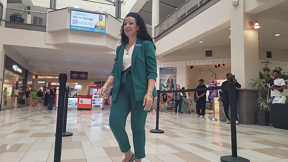 Woman in green suit, walking inside a shopping mall, smiling at camera. BGCCA