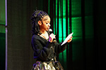 Young woman speaking into a microphone while reading from paper during an event.