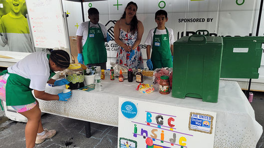 Three people stand behind a table with food, BGCCA, and the text above.