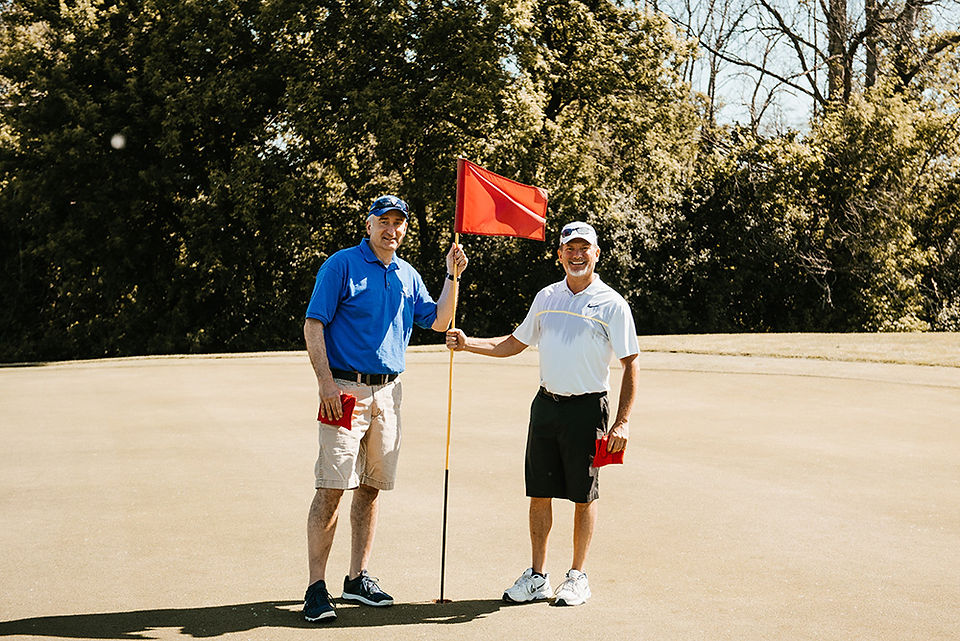 Two men pose on a golf course holding a red flag BGCCA.