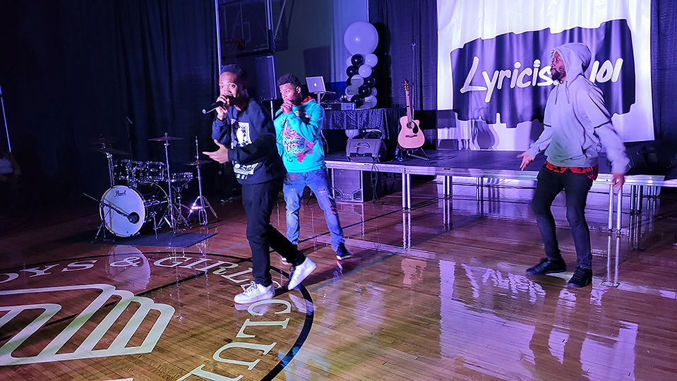 Three young men performing on stage with a banner reading Lyricist 101