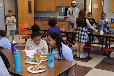 Children eating lunch and reading books at school tables, with teacher.