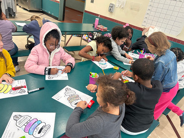 Students color at tables with drawings. Classroom setting, with drawings of shapes like BGCCA.