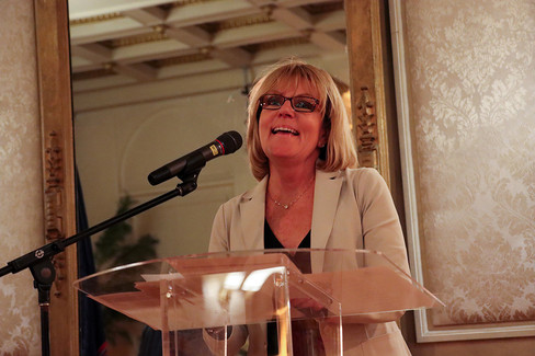 Woman speaking at podium with microphone and glasses. Background is blurry. BGCCA