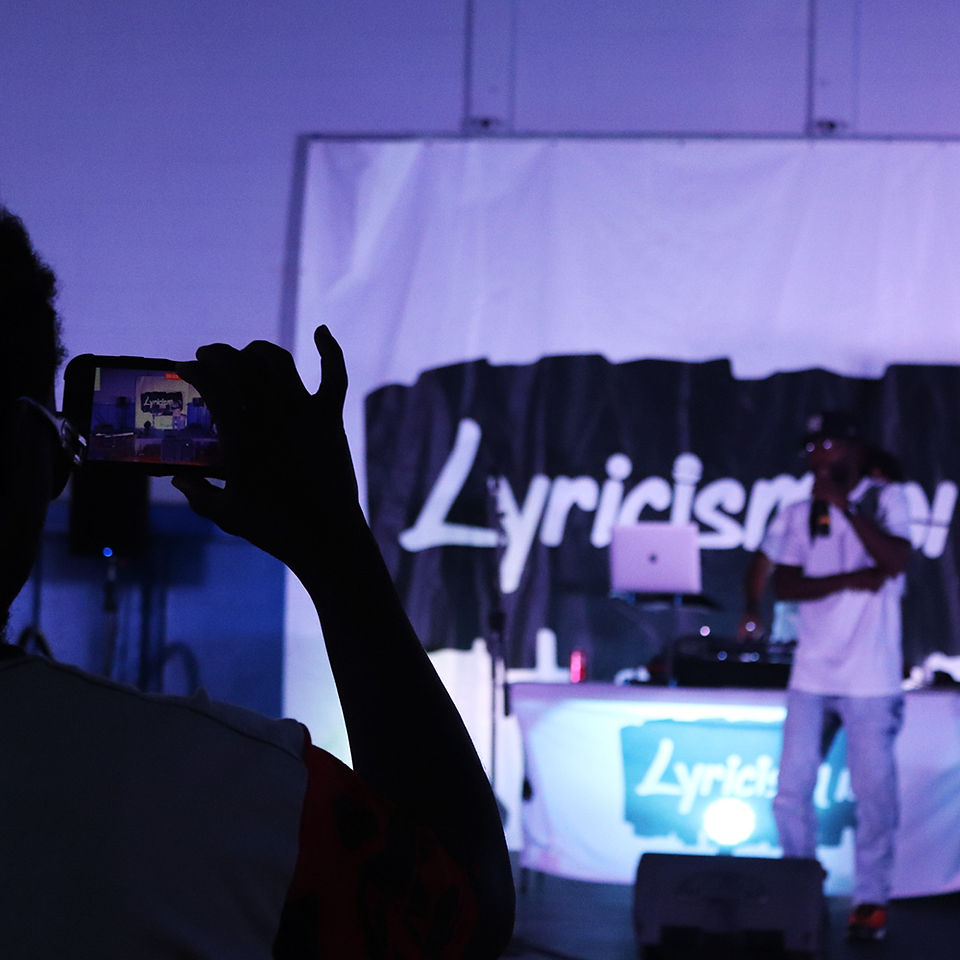 Person filming Lyricism event, DJ on stage with laptop and sound system.
