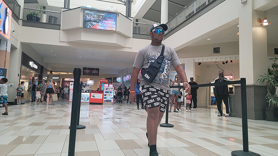 Man walking in a mall, wearing shorts and a hat, with a bag.