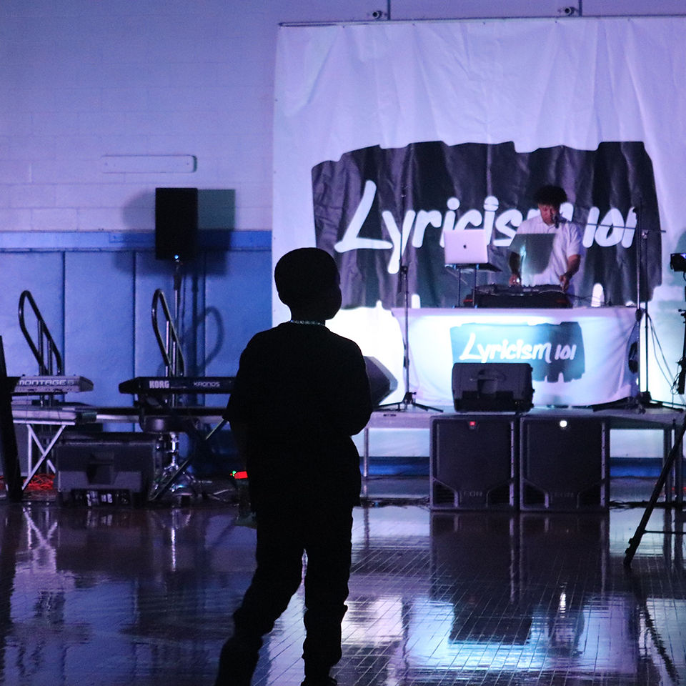Boy watches DJ with laptop behind a banner that says Lyrics 101. BGCCA