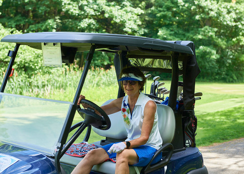 Woman driving golf cart smiling on course with golf clubs and shade.