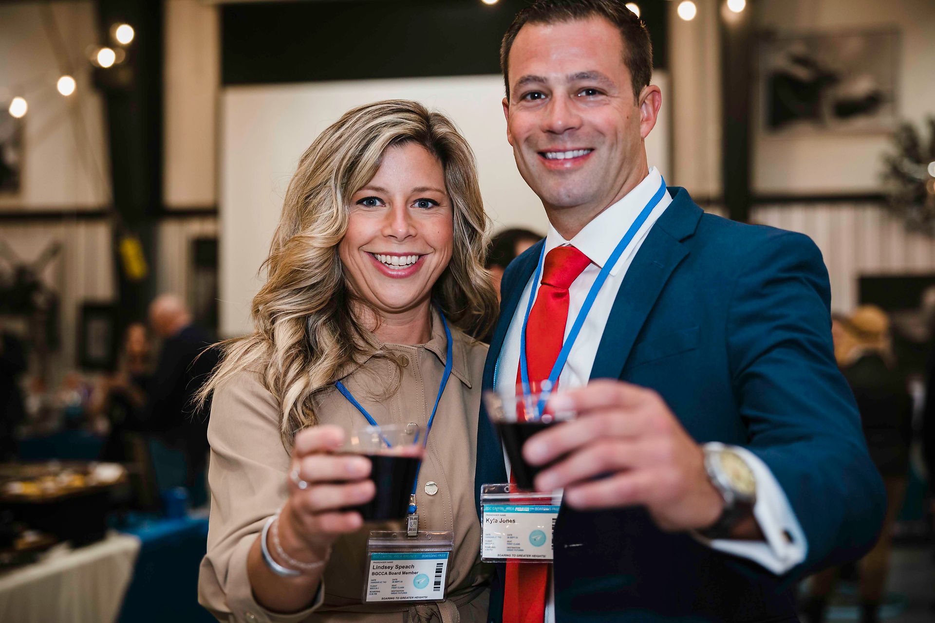 Smiling couple toasting with red wine while wearing lanyards. BGCCA