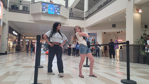 Two women posing in a mall; people and decor in background.