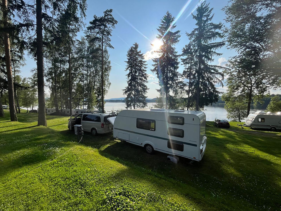 Weißer Wohnwagen mit Auto am idyllischen See zwischen Bäumen, Campingplatz in der Natur, andere Wohnwagen im Hintergrund