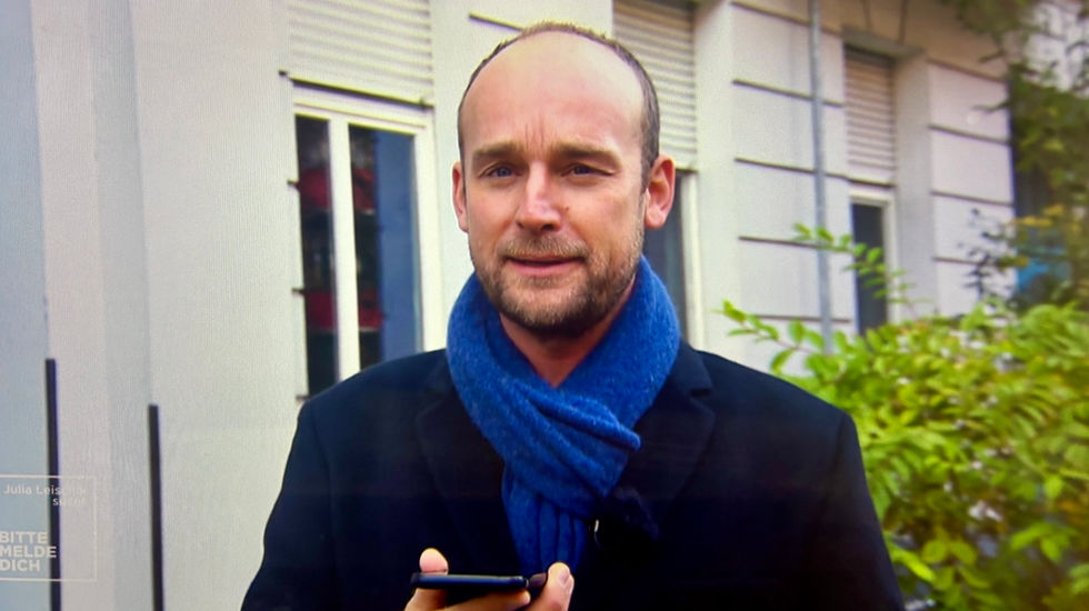 TV-Reporter Felix Schlebusch im dunklen Mantel und blauem Schal vor einem Stadtgebäude, professionell in die Kamera sprechend während eines Drehs für SAT.1.
