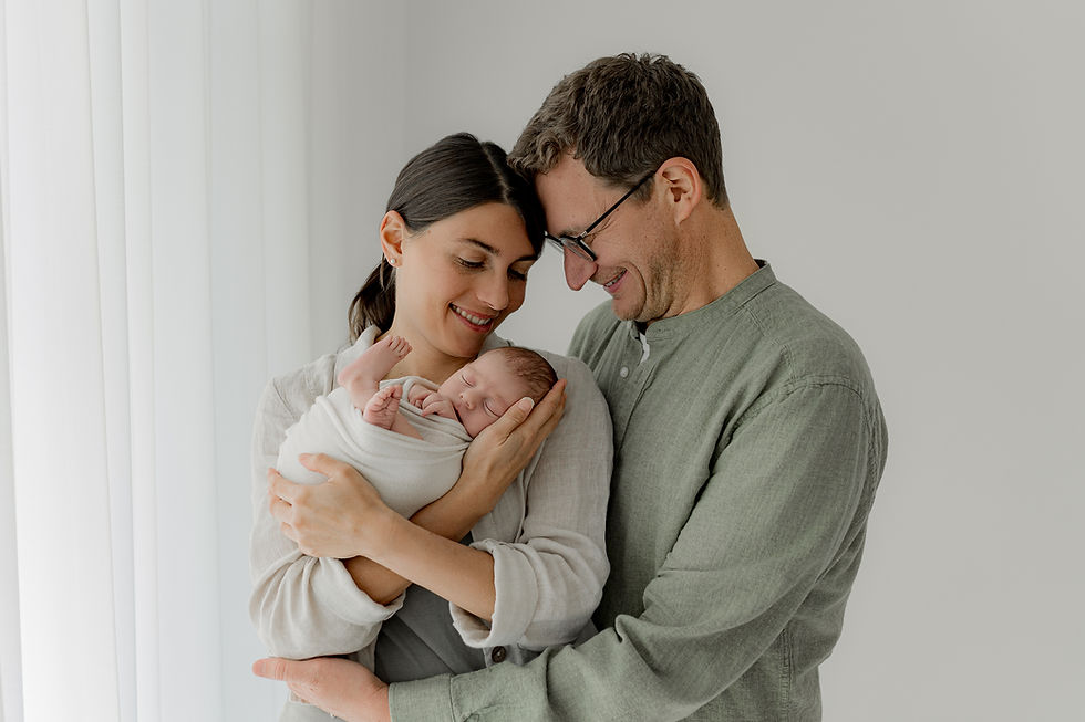 Paar hält lächelnd ihr in ein weißes Tuch gewickeltes Baby in einem hellen Raum mit weißen Vorhängen, emotionaler und liebevoller Moment.