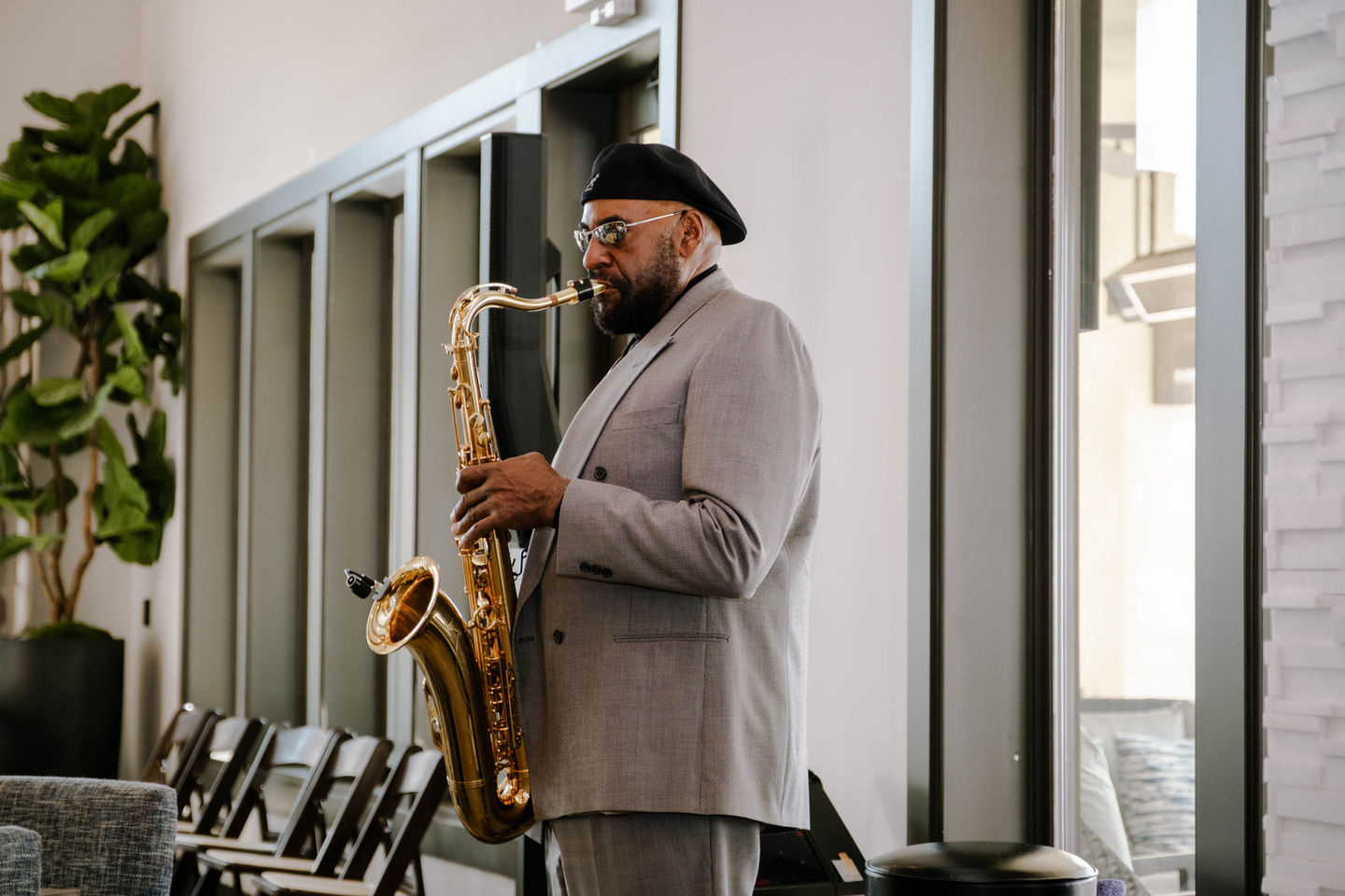 Saxophonist playing saxophone, indoor performance, wearing a gray suit, beret, and glasses.