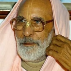 Homem idoso de óculos, barba e tilak, com pano rosa na cabeça.