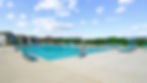Sunny outdoor pool with turquoise chairs, blue umbrellas, and a distant view of houses. Clear sky with fluffy clouds, calm and inviting.
