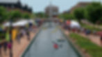 Kayakers on a canal at a lively outdoor fair with tents and colorful flags. Crowd walking along the brick paths under a clear sky.