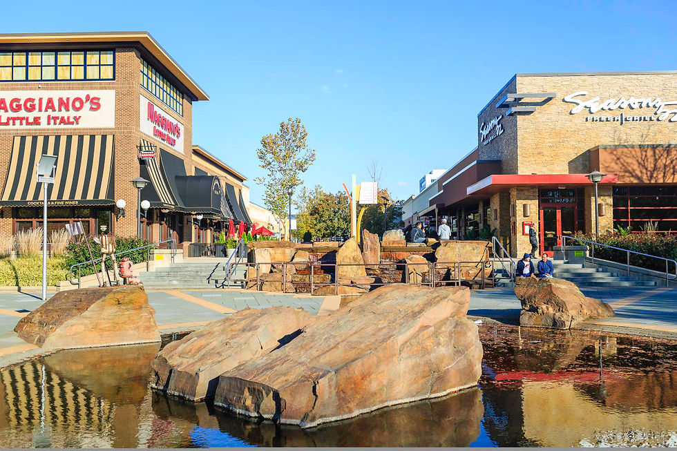 Shopping center with Maggiano's and another restaurant. People sitting near a rock fountain under a clear blue sky. Casual, relaxed mood.