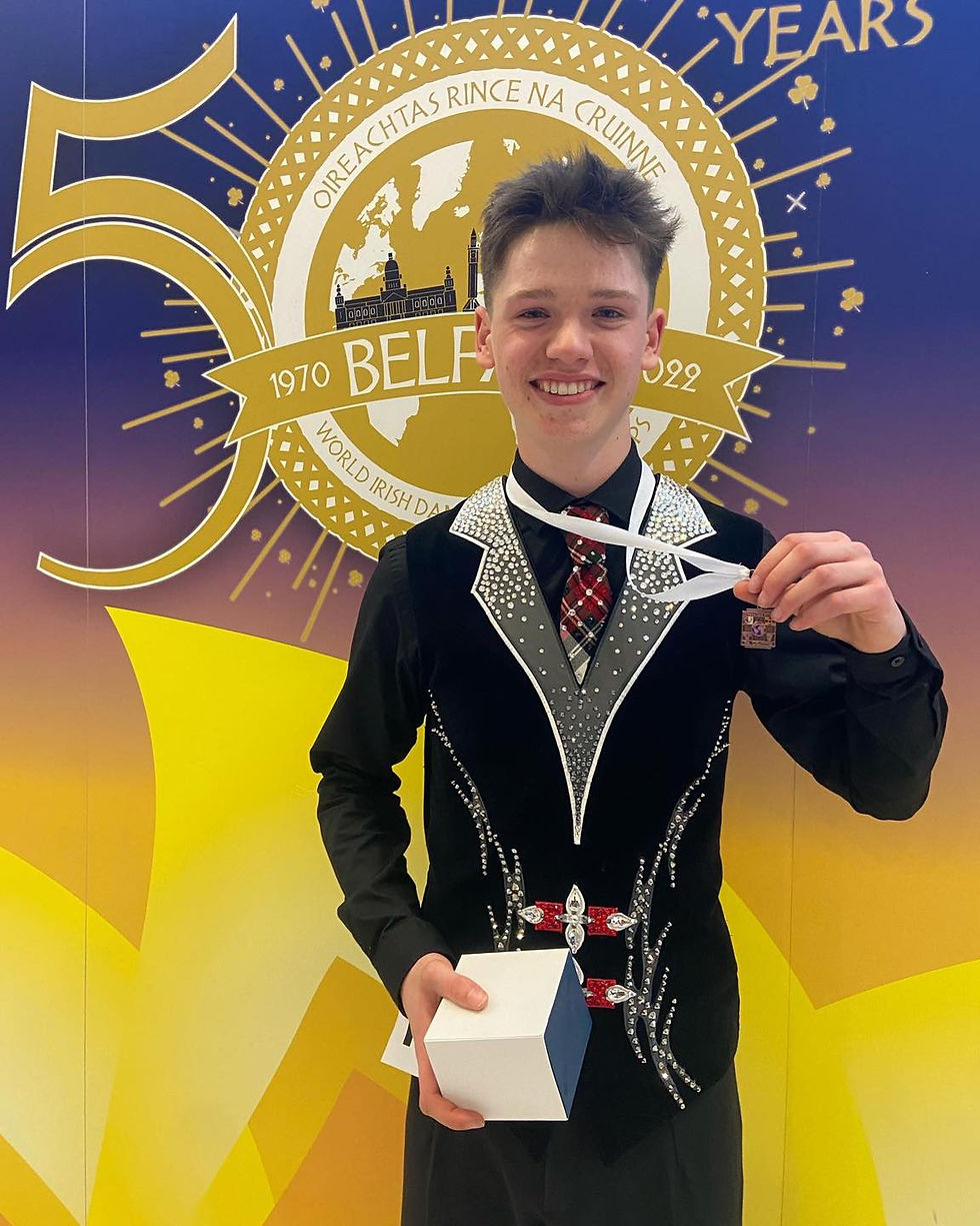 Young dancer in a studded costume holds a medal and box, smiling. Background features a 50th-anniversary banner with purple and yellow hues.