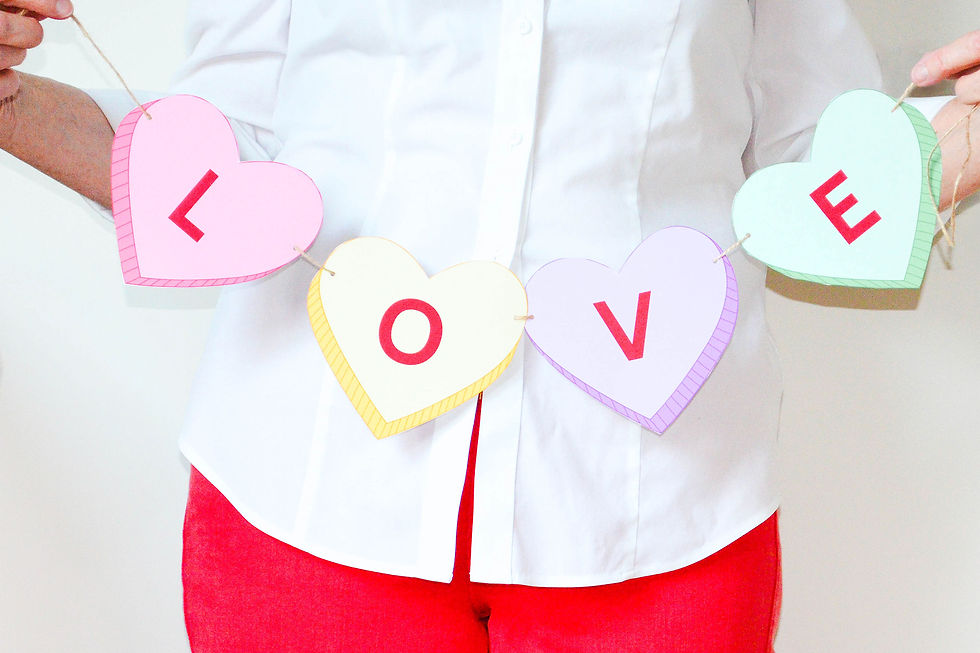 A woman with bright red pants and a white shirt holds up a banner of conversation hearts spelling the word "LOVE."