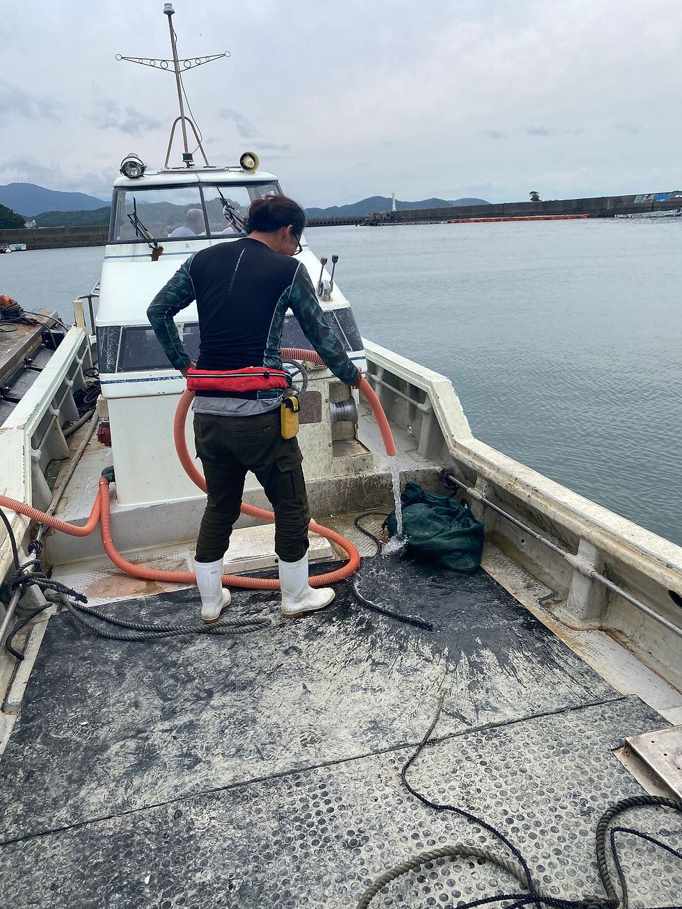 港で停泊中の小型船の甲板で、白い長靴の男性がオレンジのホースで水を流し清掃している。曇り空と静かな海が広がる。